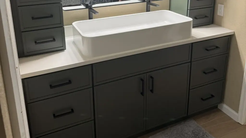 Remodeled bathroom vanity with dark gray drawers and cabinets, a white countertop, a white trough sink, and black fixtures.