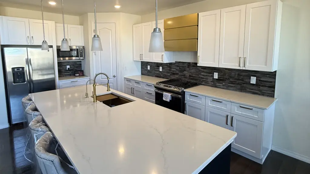 Remodeled kitchen with white shaker cabinets, a large white stone island with brass faucet, and a gray tile backsplash.